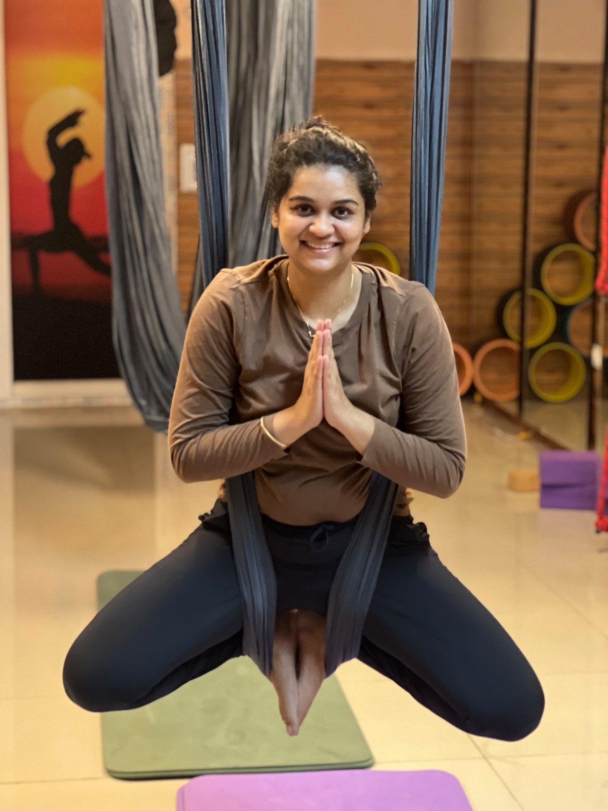 Smiling student seated in an aerial yoga hammock with hands in Namaste